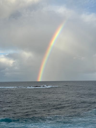 A rainbow arcs across a cloudy sky over the ocean. The ocean is a dark, choppy blue with whitecaps, and a line of rocks juts out from the water in the foreground. The scene is likely a coastal area. The location is identified as Clovelly, Australia.