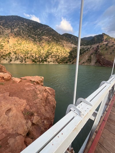 The image presents a view from what appears to be a bridge in a mountainous region, likely near or in Tabaroucht, Morocco, although this location is not explicitly confirmed by visual cues. The perspective is angled, showing a body of water, likely a river or reservoir, with a green-brown tint. The water stretches between steep, arid mountain slopes covered sparsely with vegetation. In the foreground, part of the bridge's infrastructure is visible, including white metal supports, a wooden surface, and reddish-brown rocks forming part of the terrain below. The sky above shows a mix of blue and clouds. There are no people visible in the frame. The primary subjects are the landscape elements – the water, mountains, bridge structure, and rocks.