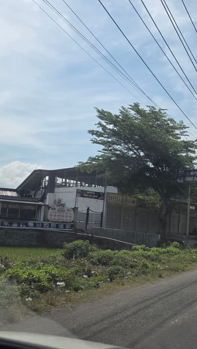 The image depicts a view of the Fans 86 Coffe establishment in Teungoh Baro, Indonesia. The building appears to be a multi-level structure with open sections and signage visible on its exterior. Adjacent to the building is a tree and a wall. A road runs alongside the structure, and utility lines are visible against a blue sky with clouds. Vegetation is also present along the road's edge. The photo seems to have been taken from a moving vehicle.
