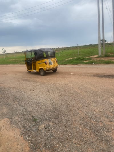 The image shows a yellow auto-rickshaw parked on a dirt road in Bulamare, Nigeria. The auto-rickshaw has a black canopy and appears to be empty. In the background, there is a field of green grass and some trees. The sky is overcast with clouds. There are some utility poles and wires visible on the left side of the image. The road surface is made of gravel and dirt. The scene appears to be set in a rural or semi-rural area. The overall lighting suggests it is daytime, likely afternoon, given the muted light and overcast conditions. There are no people visible in the image. The auto-rickshaw has some text on its side, including what appears to be a license plate that reads "AD YN". There are also some other labels and markings on the vehicle, but they are not clearly legible. The image is taken from a slightly elevated, angled perspective.