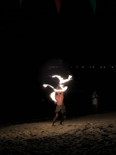 The image depicts a fire dancer performing on a beach at night. The location is Hat Khai Muk, Thailand. The fire dancer is shirtless, wearing camouflage shorts, and is wielding two flaming torches. A woman in a light-colored dress stands in the background. There are a few lights in the distance. The right side of the image shows the sand.