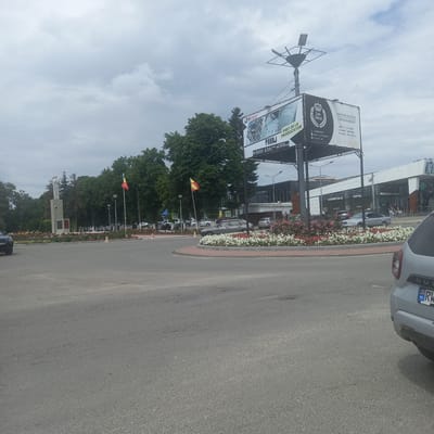 The image depicts a roundabout in Briceni, Moldova, on a cloudy day. A large billboard advertising "Lux Decor" and "IPmarket" is prominently displayed, with Romanian and Moldovan flags visible nearby. Several cars are parked or driving through the roundabout, and a monument with statues stands in the background. A building complex with shops like "Cosmetica" and "Optic" is also visible. The roundabout is adorned with vibrant flowerbeds, creating a visually appealing urban landscape.