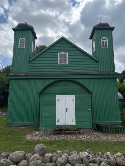 The image shows a bright green wooden mosque with two towers, located in Górka, Poland. The mosque has a central arched entrance with white double doors and a lantern hanging above. Each tower is topped with a black roof and a golden crescent moon. There are windows on the towers and on the main body of the building. The mosque is surrounded by grass, cobblestone paths, and a stone wall in the foreground. Trees are visible in the background. The sky is partly cloudy. There are no people visible in the image.