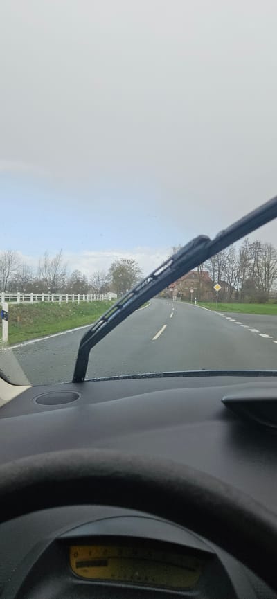 The view from inside a vehicle, likely a car, shows a wet asphalt road in Delbrück, Germany. A windshield wiper is visible in the foreground, with the road curving to the right. The road is lined with a white fence and grass. Trees are visible in the background. A road sign, possibly indicating a priority rule, is seen further down the road. Part of the car's interior, including a dashboard and speaker, is also visible. The sky is overcast.