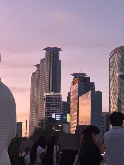 The image displays an urban cityscape at dusk in Gangnam-gu, Seoul, South Korea. The sky exhibits a gradient of light purple transitioning to a soft pink near the horizon. Dominant architectural features include a tall, multi-tiered skyscraper with a "BRUNNEN" sign on a lower section and blue and red illuminated structures at its apex. Adjacent to it, another tiered building with illuminated rooftop features is visible. To the far right, a large, curved glass high-rise building is partially visible, with letters resembling "ADELL" near its top. A digital billboard below the larger buildings displays Korean text, including "코리아나 오락실" (Koreana Game Room). The foreground contains multiple individuals, mostly viewed from the rear or in profile, positioned to observe the urban landscape. These include a person in a light-colored, textured garment on the far left, two individuals with long dark hair, and a woman in a black top on the mid-right who appears to be holding an object near her mouth. Another person in a light-colored, patterned shirt is partially visible on the far right. No explicit interactions are discernable among the individuals.
