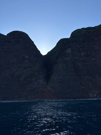 The image depicts a vast, natural coastal landscape captured during twilight hours. In the foreground, dark blue ocean water fills the lower third of the frame, exhibiting numerous ripples and reflections from a low light source. Small whitecaps are visible along the immediate shoreline. The middle ground is dominated by the base of steep, dark, rocky cliffs. The background features two massive, rugged, dark mountains rising dramatically from the sea, forming a prominent V-shaped valley or gorge in the center. Sparse green vegetation is visible near the summits of these peaks. A light source, likely the sun, is positioned directly behind the central valley, creating a bright glow against the dark silhouettes of the mountains. The sky above is clear and a pale blue, transitioning to a slightly darker shade higher in the frame. No human subjects, animals, or man-made structures are present. The scene is located in the city of Unknown, United States of America.