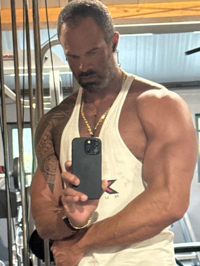 A man with a beard and short, graying hair is taking a selfie in a gym. He is wearing a white tank top, gold chain necklace, and a black watch. He has tattoos on his left arm and is flexing his right bicep. In the background, there is gym equipment, including weight racks and cables. The lighting suggests it is daytime. This image is not located in Jemelle, Belgium.