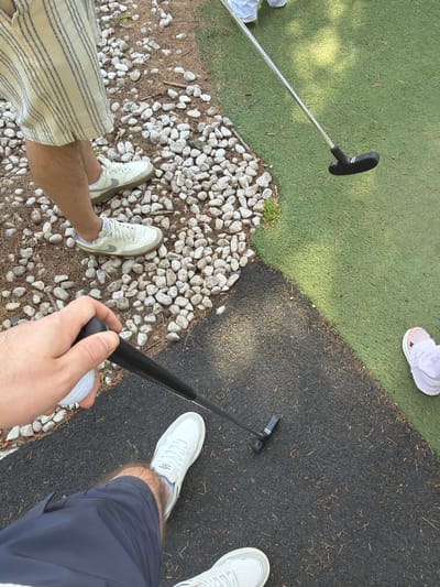 The image shows a view from above of four people playing mini golf. In the foreground, a hand holds a golf ball and putter. The person is wearing navy shorts and white sneakers. To the left, another person is wearing striped shorts and white sneakers, standing on small white rocks. To the left of that person, a golf putter lies on a green artificial turf surface. To the upper right, another person is wearing white shoes and is standing on a black artificial turf surface. A fourth person is present in the upper left corner of the image wearing pink sneakers and is also standing on the green artificial turf surface. The ground is divided into sections of artificial turf, small white rocks, and black artificial turf. The location is likely a mini golf course in Warrington, United Kingdom.