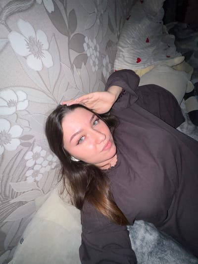 A young woman with long, dark brown hair and light green eyes lies on a bed or sofa. She is wearing a dark brown, long-sleeved garment. Her right hand is raised, touching her forehead. White wireless earbuds are visible in her ears. The background consists of floral-patterned wallpaper on the left and bedding with a striped pattern and small red hearts on the right. The lighting suggests a dimly lit environment. This scene is located in Shirkovo, Russia.
