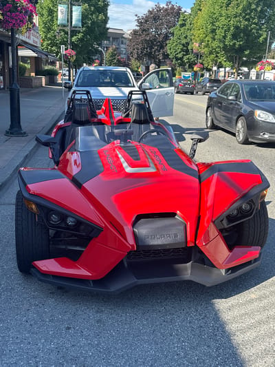 A red Polaris Slingshot, a three-wheeled autocycle, is parked on the street facing the camera in Port Coquitlam, Canada. The vehicle is bright red with black accents and has "SLINGSHOT" written vertically on the body. The street appears to be a commercial area with shops and trees lining the sidewalk. In the background, other vehicles are visible, including a white pickup truck with its door open and a dark-colored sedan. A person is partially visible through the open door of the white pickup truck. The sky is blue with some clouds. The image captures a sunny day in what appears to be a suburban or small-town setting.