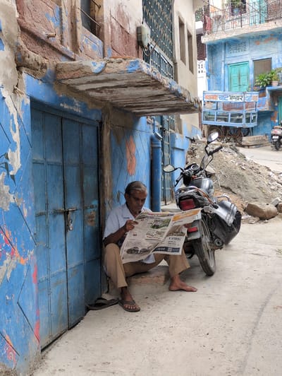 A man sits on a low step against a weathered blue door, reading a newspaper. He is wearing a light blue collared shirt and tan pants. His left foot is bare, and his right foot is clad in a dark sandal. A motorcycle is parked to his right, leaning on its kickstand. The building behind the man and the motorcycle has a textured, peeling facade with blue paint on the door and surrounding walls, and a reddish-brown upper section. In the background, a blue cart with a glass display case and large wheels is visible, as is another building painted blue with green doors. The ground is a rough, textured concrete. The scene appears to be in a narrow street or alleyway in Jodhpur, India, characterized by its blue architecture and vibrant, worn textures.