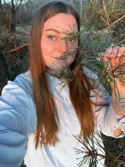 A light-skinned woman with light brown hair is pictured from a high angle. She has one visible blue-green eye and wears a light blue, collared shirt. A pine branch partially obscures her face and she holds the branch with her hand. The background appears to be woodland with visible tree trunks and ground vegetation. The exact location in Voronino, Russia, is not determinable from the image.