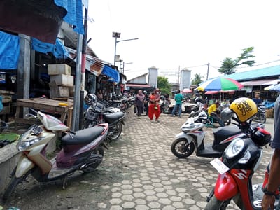 This image depicts a busy marketplace in Uruekanoe, Indonesia, with a focus on motorcycles and stalls. In the foreground, several scooters are parked on a hexagonal-patterned paved ground, some appearing dusty and slightly worn. One gold and maroon scooter is prominent on the left, while a red and black scooter with a yellow helmet on its handlebars is on the right.
The background reveals a bustling scene with numerous motorcycles, likely belonging to shoppers and vendors. People are seen walking through the market, some carrying items, while others are interacting at stalls. Several stalls are visible, covered with tarps in blue, red, and green, suggesting a variety of goods are being sold. Some stalls have corrugated metal roofs, and a wooden table with stacked boxes is also present.
In the distance, a large structure with two archways, possibly an entrance to the market or a public building, is visible. Overhead, a bright, multi-colored umbrella provides shade for a stall. The overall environment suggests a typical Indonesian market scene, characterized by vibrant activity, numerous parked vehicles, and makeshift vendor setups under a slightly overcast sky. The presence of a solar panel on a pole indicates some level of modernization within the market.