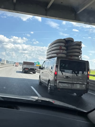 This vertically oriented image captures a bright daytime scene on a multi-lane highway, viewed from inside a moving vehicle. The most prominent subject is a silver van traveling in an adjacent lane, heavily laden with numerous light-colored, cylindrical objects, likely pipes or conduits, stacked high on its roof rack and secured with red and black straps.

The rear of the van is clearly visible, including its Russian license plate, "E762OX 198," which indicates registration in the Saint Petersburg/Leningrad Oblast region. The van's rear windows are tinted, and its taillights are illuminated. Further down the highway, a smaller flatbed truck can be seen.

The sky overhead is bright blue with scattered white, puffy clouds, suggesting clear and sunny weather. To the far left, the concrete underside of an overpass or bridge structure is partially visible. The highway itself is well-maintained with clear white lane markings. In the distance, a green landscape borders the road, and road signs with Cyrillic text can be faintly discerned, confirming the location as Russia. The overall scene depicts highway traffic under good weather conditions.