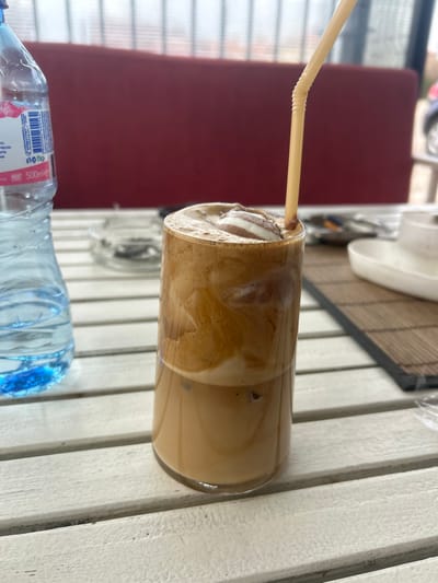 The scene, located in the city of Dolni Tsibar, Bulgaria, depicts a close-up view of a table in what appears to be an outdoor café setting. A tall, clear glass containing a layered iced coffee beverage dominates the foreground. The drink features a light brown liquid base with visible ice cubes, surmounted by a thick layer of darker brown coffee froth. A white dollop, consistent with cream or foam, is positioned on the froth, bearing a swirl of brown syrup. A beige, flexible drinking straw is inserted into the glass.

To the left of the coffee glass, a partially consumed 500ml plastic bottle of Gorna Banya water, with a blue cap and Cyrillic labeling, stands on the table. Further to the left, a circular glass ashtray containing multiple cigarette butts and ash is visible. All these items rest on a white table constructed from parallel slats. To the right of the coffee glass, a dark, rectangular woven placemat is present, holding an empty white ceramic dish. The background is blurred, revealing a red padded surface, likely seating, behind a railing or partition. Beyond this, an indistinct outdoor environment with faint hints of foliage and a vehicle is discernible. No individuals are visible in the frame.