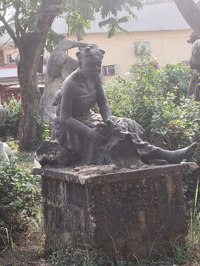 The scene depicts a weathered, dark-colored stone or concrete statue situated in a public park or garden in San Martín, Colombia. The central subject is a female figure, seated on a low structure, with her torso slightly angled and head tilted downwards. She wears her hair styled in multiple small buns or knots. Her upper body is bare, and she appears to be wearing a wrap or skirt that extends down to her lower legs. An infant or small child figure is partially visible on her back or side, attached to her form. The statue rests on a dark, rectangular concrete plinth exhibiting weathering and some organic growth.
The immediate environment around the statue features dense green foliage, including shrubs and small trees. The ground is covered with sparse grass and dirt. To the left, a large tree trunk is visible, with its branches extending overhead. In the background, a light-colored building, possibly cream or light yellow, is visible, featuring windows and an exterior air conditioning unit. Natural daylight illuminates the scene, casting soft shadows.