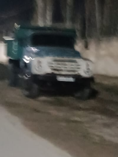 The image captures a blurry, low-light scene featuring an older model truck, likely a utility or cargo vehicle. The dominant subject is the truck, positioned slightly off-center to the left, facing towards the right foreground.

The truck's cabin and what appears to be its cargo bed or rear structure are dark teal or blue-green. The front of the truck, including the grille and headlight areas, is a lighter, almost white color. The grille features several distinct horizontal bars. Below the grille, there is a dark bumper, and in the center, a rectangular area consistent with a license plate is visible, though no characters are decipherable due to the extreme blur and low resolution. The truck has large, dark wheels, partially obscured by blur and shadow.

The entire image is heavily affected by motion blur, creating streaks of light and indistinct shapes. This blur suggests either the truck was in motion, the camera was moving during exposure, or both. Visible lights from the truck itself or ambient sources appear as elongated streaks.

The setting appears to be outdoors at night or in very dim light conditions. In the blurry background, vertical, dark, indistinct shapes can be seen, which are likely trees or poles. The ground in the foreground and mid-ground is dark and textured, resembling dirt, mud, or unpaved ground, with lighter patches on the far left that could be snow or ice. There is no clear activity depicted beyond the presence of the stationary or slowly moving truck. No discernible text is visible anywhere in the image.