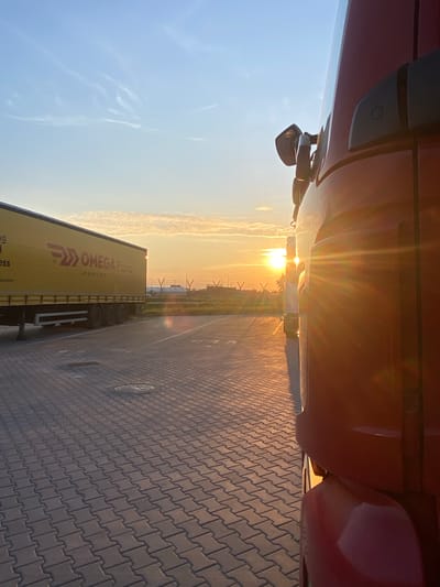 The image depicts a parking lot at sunset in Niepołomice, Poland. A red truck is parked in the foreground on the right, its side partially obscuring the view. The truck's side mirror is visible. To the left, a yellow semi-trailer truck is parked, featuring the "OMEGA Filtro LOGISTICS" logo. The ground is paved with interlocking bricks. In the background, the sun is setting, casting a warm orange glow and lens flares across the scene. The sky is a gradient of blue and orange, with some wispy clouds. Beyond the trucks, there are some industrial structures and utility poles visible. No people are present in the image.