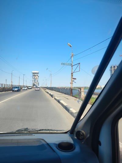 The image captures a view from inside a vehicle, looking forward along a multi-lane road on a bridge. The road is paved and shows lane markings. Several cars are driving on the road, moving away from the camera. On the left side of the road, there is a metal guardrail. On the right side, a sidewalk with a curb is visible, followed by a similar guardrail. Beyond the guardrail on the right, a body of water is visible, with buildings and construction in the distance.
A prominent feature in the distance is a large, industrial-looking metal tower structure, which appears to be part of the bridge's infrastructure. There are also numerous utility poles with wires and streetlights along the bridge. A signpost is visible on the right side of the road, with a sign indicating "р. ВОЛГА" (River Volga). The sky is clear and blue.
The image is taken in the city of Solenyi, Russia, and depicts a section of the Volga River.