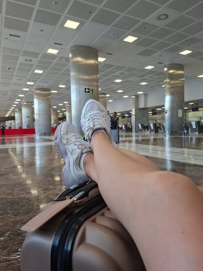 The image is a close-up shot of a person's legs and feet, wearing white sneakers, resting on a suitcase. The background appears to be an airport or a large public building with a high ceiling, large columns, and reflective floors. There are also check-in counters and signs visible in the distance. The person is positioned such that their legs and feet are the primary focus, with the suitcase in the foreground. The environment suggests a travel or waiting area. The image was taken in the city of Granadilla de Abona, Spain, which is home to Tenerife South Airport (TFS).