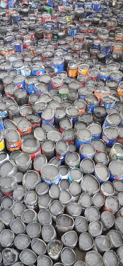 The image shows a dense, chaotic pile of discarded aluminum soda cans, many of which are crushed or dented. The cans are of various brands, including Pepsi, Fanta, and Coca-Cola, and they are covered in a layer of grey dust or dirt. Some cans are upright, while others are tilted or lying on their sides, creating a layered and uneven surface. The background is not clearly visible due to the overwhelming presence of the cans. There are no people or animals visible in the scene. The setting appears to be an outdoor area, possibly a landfill or a recycling yard, given the sheer volume of discarded cans. The lighting is somewhat diffused, suggesting an overcast day or shade. The overall impression is one of extensive waste and debris. The location in Andijani, Uzbekistan, is not discernible from the visual information present in the image itself.