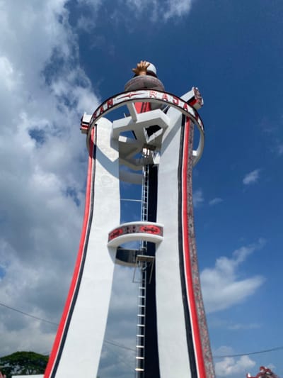 The image shows a low-angle view of a tall, white tower with red and black accents, against a bright blue sky with scattered white clouds. The tower has a conical shape, widening towards the base, and is adorned with vertical red and black stripes along its white surface. A circular band with red and white decorative patterns encircles the upper part of the tower, with what appears to be text written in red on it, reading "RAN RAJA." There is a ladder visible running vertically along the side of the tower, and a small platform with a railing extends from the tower partway up. At the very top of the tower, a golden statue of hands clasped together is visible. The background is a clear blue sky with fluffy white clouds. The scene appears to be outdoors, likely in an open area, given the unobstructed view of the sky and the presence of greenery at the bottom left of the image. This structure is identified as the Monument Simanjuntak, located in Parlondut, a village in North Sumatra, Indonesia.