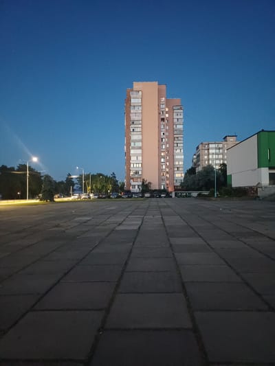An outdoor, low-angle, vertical, medium shot captures a tall, narrow, peach-colored apartment building situated in a city square. The building has numerous windows, some of which are illuminated. To the right of the main building, a shorter, grey-toned apartment building is visible. Further to the right is a white and green building with a modern design. The foreground consists of a grey, tiled square. A street with parked cars and trees is visible in the background, illuminated by streetlights. The sky above is a deep blue, suggesting it is either dusk or dawn. The scene is located in Ukrainka, Ukraine.