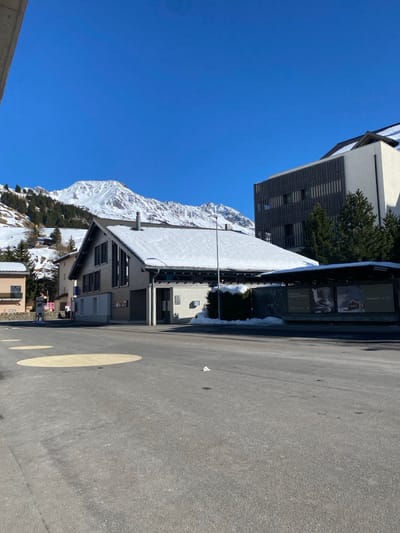 This forensic description details a daytime winter scene in Andermatt, Switzerland, under a clear, bright blue sky.
In the foreground, an asphalt road surface occupies the lower third of the image, featuring three faded yellow circular markings. A small white crumpled object rests on the asphalt near the center.
The midground is dominated by a two-story building with a steep, snow-covered gabled roof. Its lower walls are light-colored, contrasting with darker, possibly wooden, cladding on the upper story and around windows. A prominent sign above its main entrance displays "Andermatt" and additional text below it. Another sign on the building's facade shows "AND" and "SPORT." To the right of this building, a covered bus stop shelter is visible, featuring several advertising posters behind its transparent panels. A tall, dark streetlight pole stands between the building and the shelter.
To the left of the central building, another light-colored building with dark roof sections is partially visible. An indistinct, solitary figure is faintly discernible near the base of this building on the far left.
Behind the central building and bus shelter, a larger, modern structure with a multi-tiered roof and a combination of white facades and dark slatted cladding is present.
The background is dominated by a range of snow-capped mountains, partially covered with evergreen trees, under the cloudless sky. Patches of accumulated snow are visible on the ground around the buildings and at the base of the bus shelter. No explicit actions or interactions are discernible among the sparse human presence.