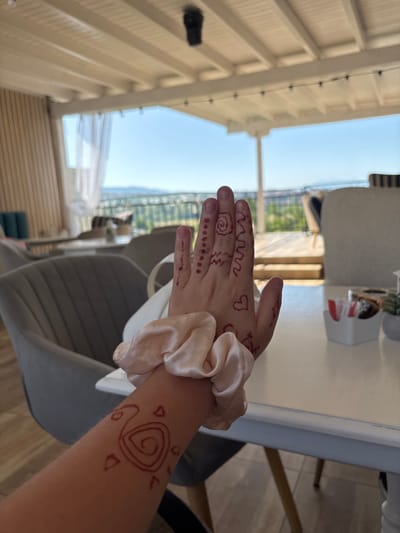 A person's arm and hand, adorned with red henna designs, are held up in the foreground of the image. The henna on the hand depicts a spiral, zig-zag lines, and a heart shape. On the forearm, a swirling spiral design is visible, along with small triangular shapes. A light-colored scrunchie is worn around the wrist. The background suggests an outdoor seating area of a restaurant or cafe, with tables and chairs visible. Beyond this, a panoramic view of a city or town is seen, with rolling hills and a clear blue sky. The setting appears to be a resort in Ognyanovo, Bulgaria. The overall lighting indicates daylight, and the environment seems relaxed and leisurely.