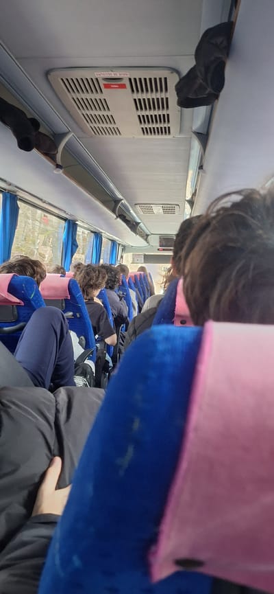 The image depicts the interior of a passenger bus or coach, specifically an aisle view looking forward from a seated position, reportedly within the city of Tsar Kaloyan, Bulgaria. Approximately ten to fifteen individuals, primarily young adults or teenagers, are seated in blue upholstered seats with pink headrest covers. Passengers are seen from behind, exhibiting various casual attire including dark jackets, t-shirts, and pants. One individual in the immediate foreground, positioned to the right, has dark, curly hair. Another passenger in the left foreground is wearing dark blue pants and a dark jacket, with their leg extended into the aisle area.
Overhead, a light gray ceiling features an air vent or light fixture with a red and white label displaying "ISSUE DE SECOURS" (Emergency Exit) and "TIRER" (Pull) in French. Overhead luggage compartments line both sides of the bus. Blue curtains partially cover the windows on the left, through which a blurred, tree-lined exterior is visible, suggesting the bus is in motion. A small screen or monitor is mounted on the ceiling towards the front of the vehicle. A dark, folded garment or object hangs from an overhead rack on the right side. No active interactions between passengers are discernible; all appear to be passively seated during travel. The person taking the photograph has a hand visible in the lower left, resting on their dark attire.