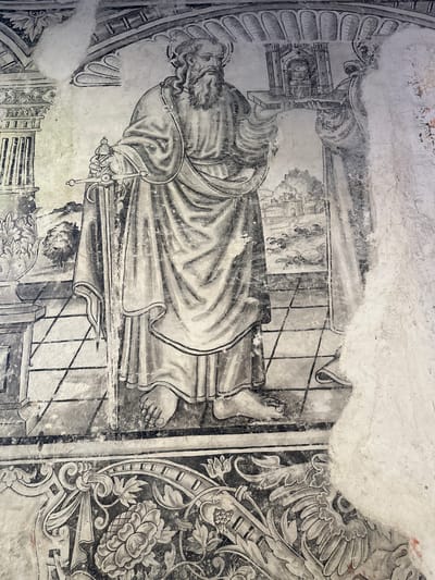 The image is a photograph of a faded fresco on a wall in Huejotzingo, Mexico. The fresco depicts a male figure with a beard, wearing a toga and sandals. He holds a sword in his left hand and a miniature architectural structure in his right. The figure stands on a patterned floor within a shallow architectural setting, indicated by columns and arches. A second, partially obscured figure is visible to the right. In the background, a landscape with buildings and vegetation is depicted. The lower portion of the fresco features an ornamental border with floral motifs. The fresco is in a state of deterioration, with large areas of the plaster missing, revealing the underlying wall. The colors are monochromatic, rendered in shades of gray and black.