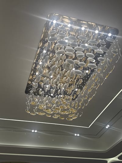 This image shows a modern, rectangular chandelier made of numerous oval-shaped, clear and golden elements suspended from thin wires. The chandelier is illuminated by embedded lights, which create a bright and reflective effect, casting streaks of light onto the surrounding wall and ceiling. The surrounding environment appears to be an interior space with smooth, neutral-colored walls and ceiling, possibly a modern home or commercial building. There are also some subtle linear lighting elements integrated into the ceiling design. The overall impression is one of contemporary elegance and sophisticated lighting design. There is no visible text in the image. The time of day and weather are not discernible from the interior setting. The specific location within Jezqazğan, Kazakhstan is not identifiable from the visual information.