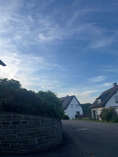 This image depicts a residential street in the city of Wiehl, Germany. The scene is captured on a clear day with a blue sky dotted with wispy white clouds. On the left side of the image, a curved, dark stone wall lined with dense green foliage forms the foreground. Beyond the wall, a two-story white house with a dark tiled roof, solar panels, and several windows is visible. Further down the street, another white house with a similar dark tiled roof and a dormer window is present, partially obscured by bushes and flowering plants. The street itself is paved and appears to be sloping upwards. Hills covered in trees are visible in the background. The overall atmosphere is tranquil and suburban.
