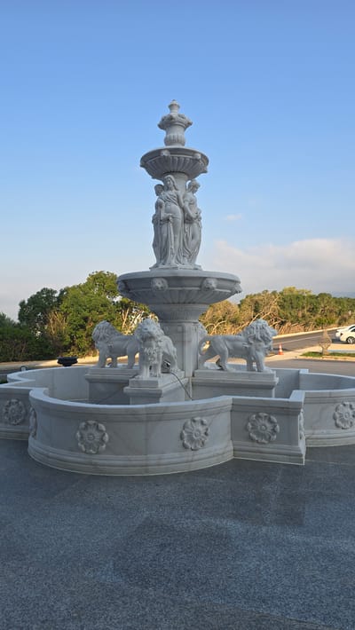 An ornate white marble fountain stands in the foreground, featuring multiple tiers. The topmost tier is a decorative finial. Below this, a large basin is adorned with carved lion heads. Supported by figures of women, the main body of the fountain rises from a circular base. Surrounding the central structure is a lower, curved basin with decorative floral carvings. Several sculpted lions stand guard at the base of the fountain, positioned on individual plinths. The fountain is situated on a grey, speckled paved area. In the background, lush green trees line a gentle slope leading up to a road, where a white car is parked. The sky above is a clear, pale blue with a few wispy clouds. The scene suggests an outdoor setting, likely a public space or a well-maintained garden area in Agios Epiktitos, Northern Cyprus.