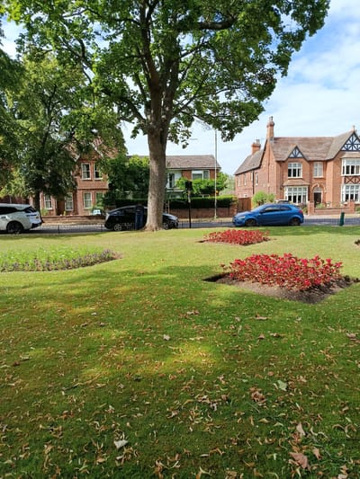 A tranquil daytime scene unfolds in Bedford, United Kingdom, featuring a well-maintained park with lush green grass. Scattered fallen leaves suggest the season might be autumn. In the foreground, two neatly arranged flower beds showcase vibrant red blooms, adding a splash of color to the landscape. Beyond the grass, a dark metal fence delineates the park from a paved road.
Several cars are parked along the road, including a white SUV, a dark hatchback, and a bright blue hatchback. A lone person stands near the dark hatchback, seemingly engaged with their phone. In the background, traditional brick houses with tiled roofs line the street, some featuring decorative gables and white-framed windows. Large, mature trees with abundant green foliage dominate the left side of the frame, casting dappled shadows on the grass. The sky above is a mix of blue and white clouds, indicating fair weather. The overall atmosphere is peaceful and serene, capturing a typical day in a suburban or parkland setting. No visible text is present in the image.