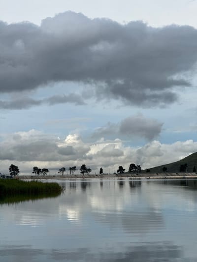 The image depicts a body of water, likely a reservoir or canal, in Ust-Dzheguta, Russia. The water's surface is relatively calm, reflecting the overcast sky and a line of trees and a concrete embankment visible on the opposite shore. The sky is filled with dramatic, dark grey clouds in the upper portion of the frame, transitioning to lighter grey and white clouds towards the horizon. A green, grassy bank with some taller reeds is visible on the left side of the frame, extending into the water. Beyond the water and embankment, a sloping, grassy hill is visible on the right, dotted with a few trees. The scene appears to be captured on an overcast day, creating a muted and somewhat somber atmosphere. No people or specific actions are discernible in the image.