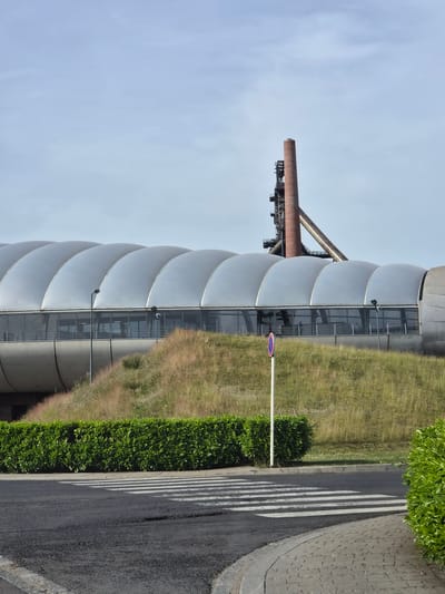 The image shows an exterior view of the Blast Furnace Belval in Sanem, Luxembourg. The building is characterized by its unique, bulbous silver exterior. A tall, rusty smokestack extends beyond the building's roofline. Vegetation, including grasses and bushes, lines the side of the building, bordering a paved road with a crosswalk. A sign with a red and white symbol is positioned along the road. The sky is a pale blue.