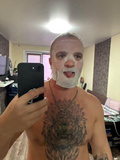 In Murmansk, Russia, a man with short, graying hair is taking a selfie in a room. He is wearing a white sheet face mask that covers his forehead, eyes, nose, and mouth, with his tongue sticking out through a cutout. He has a large, intricate tattoo of a lion's head on his chest and shoulder. He is wearing a silver chain necklace. The man is holding a black smartphone in his left hand. The background shows a wall with striped wallpaper, a window with pink curtains, and some furniture, including a television set and a bed with striped bedding. The lighting suggests an indoor setting with overhead illumination.