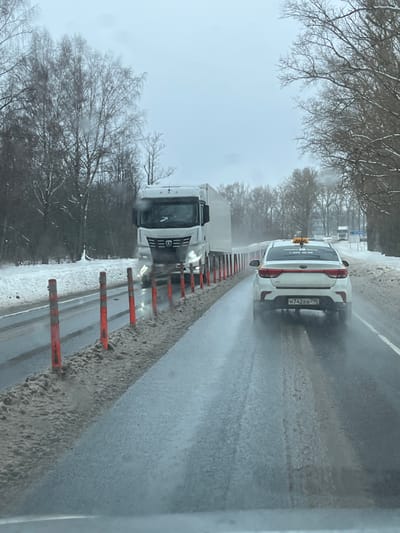 The image presents a wet, multi-lane highway scene in Podmosh'ye, Russia, viewed from inside a vehicle under an overcast sky. The road surface is dark, reflective asphalt, covered in a thin layer of water or slush. Snow banks line both sides of the road, and a significant accumulation of dirty snow and slush separates the leftmost lane from the right via a series of orange and black flexible bollards.

A white KAMAZ semi-truck with a box trailer is in the leftmost lane, approaching the viewer with its headlights illuminated. In the right lane, ahead of the viewer's position and moving away, is a white Kia Rio sedan functioning as a Yandex Taxi, identified by a yellow roof-mounted sign with Cyrillic text "Яндекс Такси" and a phone number. The taxi's rear license plate reads H 742 BB 799 (Russian Federation). A second white truck is visible in the distant right lane. Bare deciduous trees line the road on both sides. A white road sign on the right shoulder in the distance indicates "Подомошье 2 км" (Podmosh'ye 2 km). No individuals are distinctly visible.