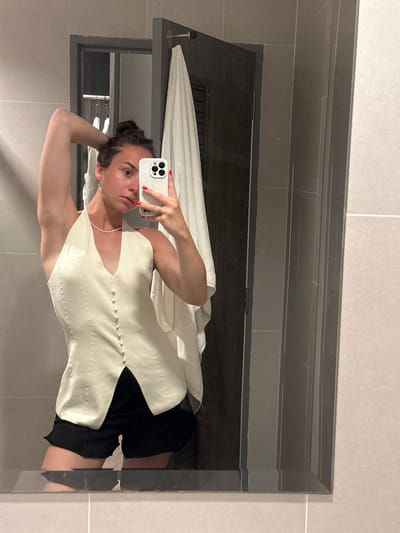A woman takes a selfie in a mirror in a bathroom. The woman has dark hair pulled back in a bun and is wearing a cream-colored, sleeveless top with buttons down the front and black shorts. The bathroom has gray tile walls. A dark wood shelf and a white towel are visible in the mirror behind the woman. The location is Baan Klong Prao, Thailand.