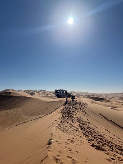The image displays a vast desert landscape dominated by numerous orange-brown sand dunes under a clear, brilliant blue sky, consistent with the terrain surrounding Rissani, Morocco. The sun is high in the sky, visible as a bright, overexposed point with lens flares in the upper center. In the midground, a white four-wheel-drive vehicle, equipped with a roof rack, is parked on a relatively flat expanse of sand between dunes. Two individuals are present near the vehicle: one person, dressed in light-colored clothing, stands upright, facing the rear of the vehicle; another person is crouched on the sand to the right of the standing individual, with their back to the viewer. The foreground features a prominent sand dune ridge extending from the lower left towards the midground, clearly marked with numerous footprints. The background consists of rolling sand dunes extending to the horizon.