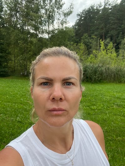 The image presents a close-up, chest-up outdoor portrait of an adult female located in Strigailiškis, Lithuania. The subject, a female with light-colored hair pulled back, blue eyes, and a neutral facial expression, looks directly forward. She wears a white, ribbed tank top, and a thin gold-colored necklace with a small circular or star-shaped pendant is visible on her chest. A streak of white substance or residue is present on her left and right jawline, extending onto the chin. The immediate background consists of a well-maintained, lush green grass field. Beyond the field, a dense line of various trees and vegetation is visible under an overcast, light grey sky. The left side of the background features deciduous trees with lighter green leaves, while the right displays darker coniferous trees and a mix of reeds and shrubs.