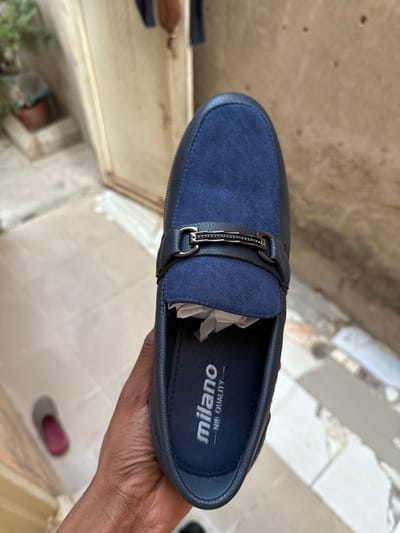 A close-up shot features a single dark blue loafer-style shoe being held by a person's hand, positioned against a softly blurred background. The shoe is primarily dark blue, with a distinct section of contrasting material, possibly suede or textured fabric, covering the vamp. A decorative silver-toned metal buckle with an intricate pattern is prominently placed on the shoe's upper, bridging the main body and the vamp.
Inside the shoe, white tissue paper is visible, suggesting it is a new item. The insole displays white text, oriented upside down from the viewer's perspective, reading "MILANO" in a larger font and "NIP QUALITY" below it. The hand holding the shoe has a dark skin tone. The background, though out of focus, reveals light-colored tiled flooring and architectural elements, hinting at an indoor or semi-outdoor setting. The bright, diffused lighting indicates it is daytime. The scene appears to be set in a location consistent with Dan Maliki, Nigeria, where the shoe is being presented or inspected.