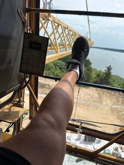 This image shows a first-person perspective from inside a crane cabin, looking out. The subjects are a person's leg and foot, wearing a black shoe and white socks, positioned on the edge of the cabin. The background reveals a construction site with a large yellow crane arm extending upwards and to the left. Beyond the construction site, a body of water, possibly a lake, is visible, with trees lining the far shore and a hazy sky above. The scene appears to be outdoors during the daytime, with sunlight casting shadows within the cabin.
A notable object in the foreground is a control panel or data plate with various measurements and labels. Text on this plate is in Polish and English, including "ODLEGŁOŚC WOZKA" (Trolley Distance), "Ausladung Radius Portée" (Outreach Radius), and "Tragfähigkeit Capacity Charge" (Load Capacity). Specific load capacities in kilograms are listed against corresponding outreach radii in meters. The time of day is likely late morning or early afternoon, given the bright sunlight. The weather appears to be clear. The location context of Mikołajki, Poland, suggests this crane is operating near a lake or waterfront area. The image conveys a sense of being high up and potentially involved in construction or maintenance work.