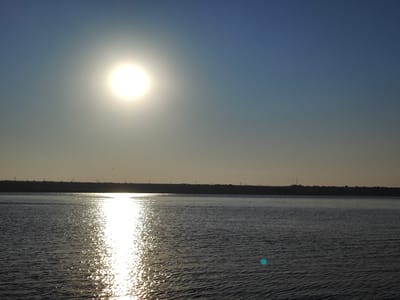 The image shows a wide shot of a body of water, likely a river or lake, under a clear sky. The sun is high and bright, casting a strong reflection on the water's surface, creating a shimmering path of light. In the distance, a dark, silhouetted landmass forms the horizon, with a faint line of indistinct structures, possibly power lines or buildings, visible along its edge. The water itself is dark and appears to have small ripples. The sky transitions from a bright blue at the top to a lighter, hazy tone near the horizon, where it meets the land. There are no visible people or specific landmarks that would definitively place this image within Nezavertaylovka, Moldova, beyond the provided context. The overall scene is serene and natural, dominated by the sun and its reflection.