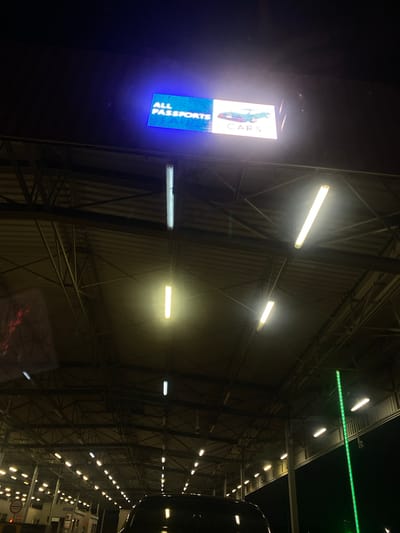 The image shows the interior of a large, industrial building, possibly a warehouse or exhibition hall, at night. The setting is dimly lit, with the primary light sources being numerous fluorescent tube lights running along the ceiling and walls, and a brightly lit digital sign on the left. The digital sign displays text and graphics, with the word "CARS" prominently visible in a vertical orientation, along with what appears to be car imagery. Below this, another blue digital display shows a pattern of white lights, possibly indicating a number or status.
The structure of the building is characterized by exposed metal beams and corrugated metal siding on the walls. Reflections are visible on the floor, suggesting it is a smooth, possibly polished surface, and hints at the presence of vehicles or other objects within the space. The overall mood is utilitarian and somewhat stark, with the artificial lighting creating long shadows and highlighting the architectural details. The time of day is clearly night due to the darkness outside and the reliance on artificial illumination. No people are visible in the frame, focusing the attention on the architecture and the illuminated displays. The location context of Terespol, Poland, suggests this might be a car dealership, exhibition center, or a similar commercial or industrial facility in that area.