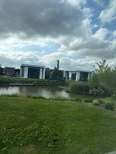 The image presents a view in Berkhout, Netherlands, featuring modern architecture alongside a natural landscape. Two contemporary, white, minimalist structures with black-framed windows are situated near a waterway. One structure is partially obscured by foliage. The water reflects the sky and the buildings. Lush green grass, interspersed with small yellow flowers, covers the foreground. Above, a cloudy sky with patches of blue forms the backdrop. No people are visible.