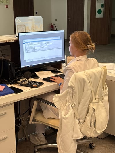 A blonde woman wearing a white lab coat is sitting at a reception desk, facing a computer monitor. The monitor displays a complex interface with various fields and input boxes, suggesting she is operating a software system, possibly for data entry or patient management. A calculator, a smartphone, and a blue notebook are on the desk in front of her. A white quilted jacket is draped over the back of her chair. The desk is white, and to the left, a black computer tower is visible. In the background, there are wooden doors and a fire extinguisher mounted on the wall. The environment appears to be an indoor office or administrative space. The sign on the desk reads "КАССА", meaning "cashier" or "checkout" in Russian.