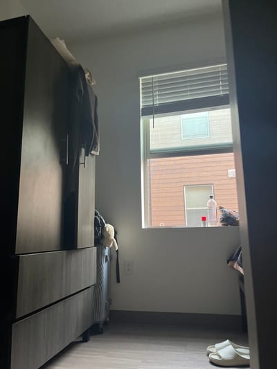 In a bedroom in Snyderville, United States of America, a tall, dark wood wardrobe stands to the left of the frame. A brown sweater is draped over the top of the wardrobe, with a light-colored garment visible behind it. To the right of the wardrobe, a suitcase with a textured surface is placed on the floor, and a light-colored stuffed animal is attached to the suitcase's handle. A window with horizontal blinds is situated in the center of the back wall, revealing an exterior view of buildings with horizontally arranged wood siding and a smaller window. On the windowsill, a white plastic bottle, a small red container, and a dark-colored bag with a patterned fabric are present. A white electrical outlet is visible on the wall to the right of the suitcase. In the foreground, on the right side of the image, a pair of white slides are on the floor. The floor is covered with light-colored wooden planks. The lighting suggests it is daytime.