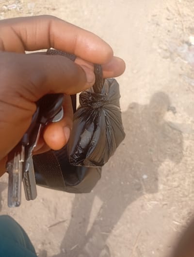 A person is holding a black plastic bag, tied at the top. The person is holding a key in their other hand. The image is taken from a low angle and the person is standing on what appears to be a dirt road or path. The background is blurred and shows a dry, sandy environment. The coordinates provided are for a location in the city of Assai Sabongida, Nigeria. The scene is located on a dusty, outdoor path. There is a person's arm and hand visible in the image. The hand is holding a black plastic bag, tied off at the top. The bag is empty. A set of keys is partially visible, being held in the same hand, next to the plastic bag. The person is wearing a blue shirt, but most of it is not in the image. The path is a dirt path and has a rough, dry texture, appearing to be a common pedestrian path. There is a shadow visible of the person's body in the path, indicating that the person is standing in bright, natural lighting. The scene is an unassuming, typical, rural setting, with no specific details indicating anything more than everyday life.